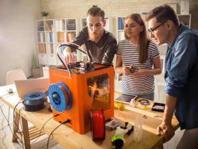 Three young engineers standing at wooden desk and using 3D printer in order make small prototypes, interior of modern laboratory on background
