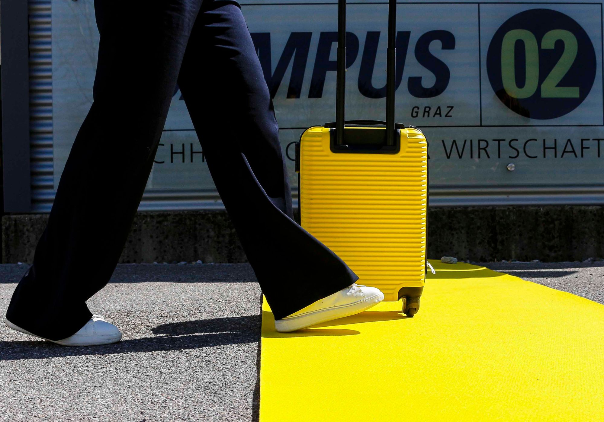 Person mit gelben Koffer schreitet über einen gelben Teppich vor der FH Campus 02